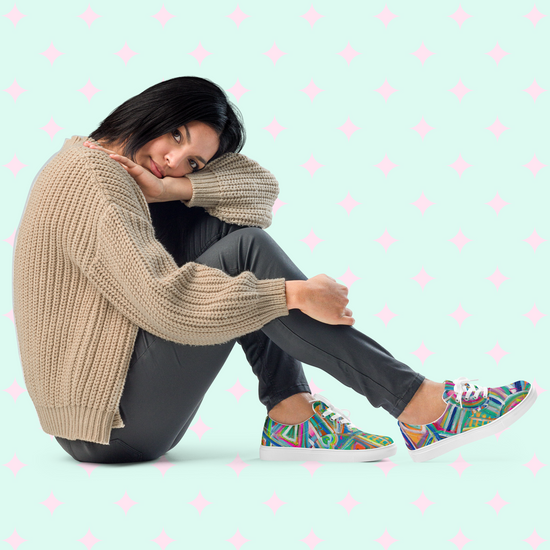 A person with shoulder-length hair sits against a light blue backdrop with pink diamond patterns. They wear a cozy beige sweater, dark pants, and vibrant Geometric Rainbow Canvas Sneakers by My Favourite Colour is Rainbow, gazing thoughtfully to the side.