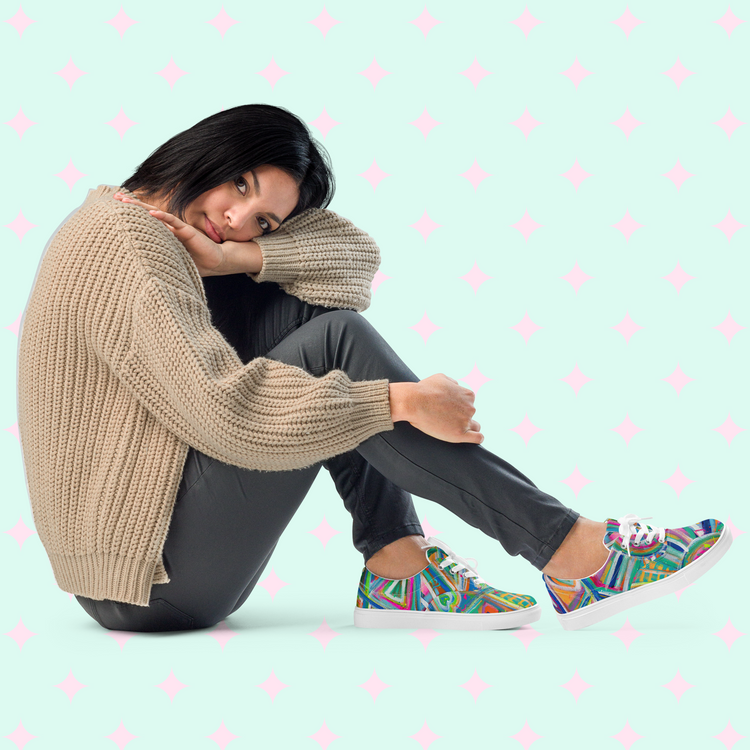 A person with shoulder-length hair sits against a light blue backdrop with pink diamond patterns. They wear a cozy beige sweater, dark pants, and vibrant Geometric Rainbow Canvas Sneakers by My Favourite Colour is Rainbow, gazing thoughtfully to the side.