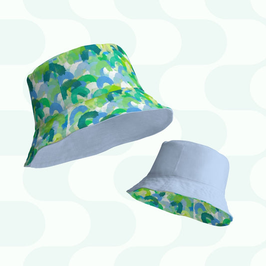 Against a geometric pale background, two bucket hats by My Favourite Colour is Rainbow are showcased. The larger one boasts a colorful green, blue, and white pattern. The smaller light blue hat has a band of the same vibrant design on its brim. Perfect for achieving that Style BFF.