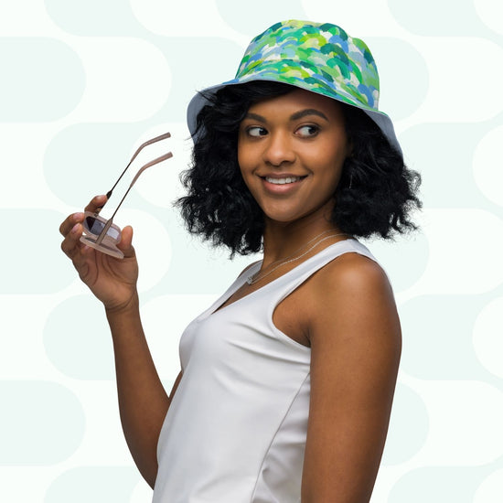 A woman with curly hair wears a Green Rainbow Reversible Bucket Hat from My Favourite Colour is Rainbow, radiating vibrant style. She pairs it with a white sleeveless top and holds glasses, smiling to the side against a light background with abstract patterns.