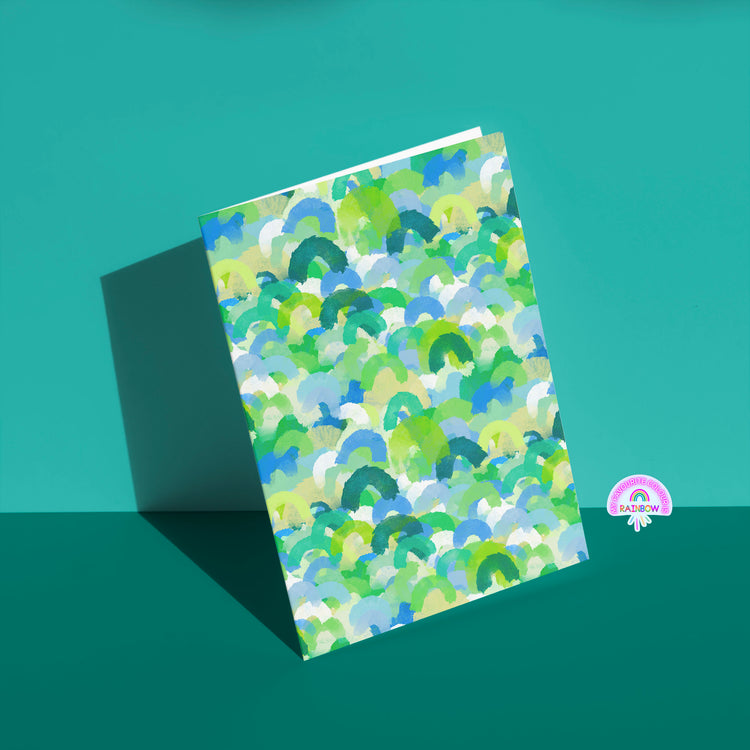 The Green Rainbow greeting card from My Store features vibrant blue, green, and yellow brushstrokes against a teal backdrop, perfect for birthdays or special occasions. It includes a small logo with a rainbow in the bottom right corner.