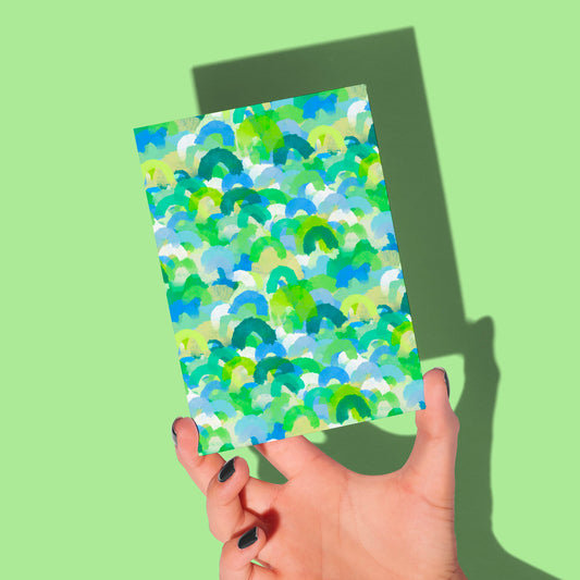 A manicured hand holds a Green Rainbow Greeting Card by My Store over a green background. The card features abstract blue, green, and white brushstrokes in a vibrant design. A shadow of the hand and card is visible.