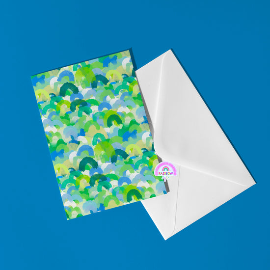 The Green Rainbow Greeting Card by My Store, ideal for birthdays, features lively green, blue, and yellow brushstrokes on a white envelope decorated with a RAINBOW sticker and rainbow icon against a blue backdrop, capturing the essence of color celebration.