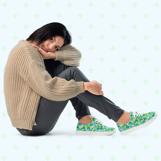 A woman with short black hair sits on the floor, head resting on one knee. She's in a beige sweater and dark pants, paired with My Favourite Colour is Rainbow's Green Rainbow Canvas Sneakers, featuring green and blue rainbow patterns.