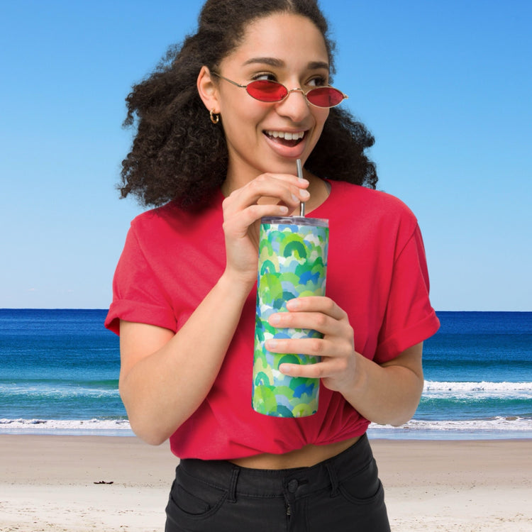 A woman with curly hair, wearing red sunglasses and a red top, sips from a My Favourite Colour is Rainbows Green Rainbow Stainless Steel Tumbler with a metal straw. Shes in front of a beach scene featuring a clear blue sky and ocean waves.