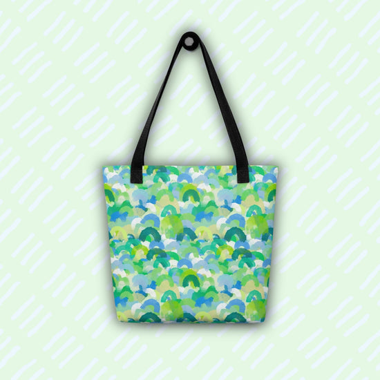The Green Rainbow Tote Bag by My Favourite Colour is Rainbow hangs against a light green background with diagonal white stripes. This colourful tote features black handles and a green and blue watercolour rainbow design.