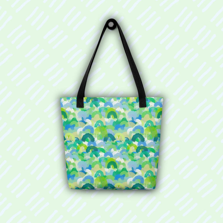 The Green Rainbow Tote Bag by My Favourite Colour is Rainbow hangs against a light green background with diagonal white stripes. This colourful tote features black handles and a green and blue watercolour rainbow design.