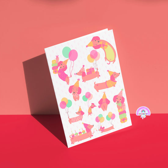 The Happy Hotdogs Birthday Card by My Store features colorful dachshunds in party hats with balloons and presents, standing open against a pink background. It includes a rainbow logo in the bottom right corner.