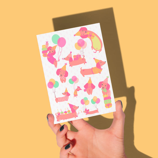 A hand holds My Stores Happy Hotdogs Birthday Card against a yellow background, showcasing playful dachshunds in party hats with balloons and gifts, while the shadows of the hand and card add depth to the joyful scene.