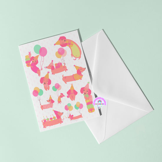 The Happy Hotdogs Birthday Card by My Store rests on a green surface, featuring fun dachshunds in party hats with balloons. Accompanied by a white envelope, the design pops with vibrant pink, yellow, and teal.
