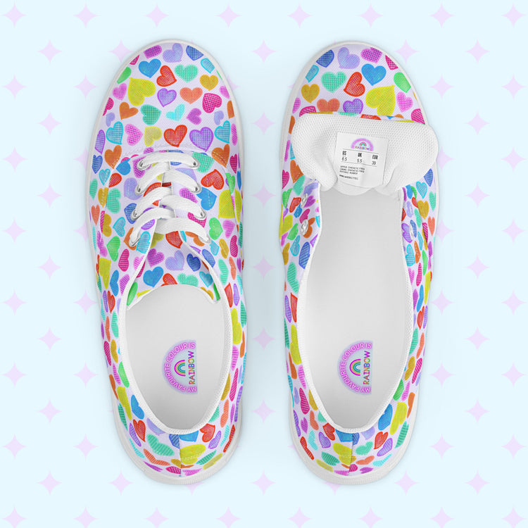 A top-down view showcases a pair of Heart Attack Canvas Sneakers by My Favourite Colour is Rainbow, each featuring heart patterns of various colours. The left shoe is laced, while the right shows size labels on the exposed tongue.