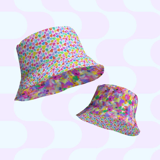 Two vibrant reversible bucket hats, the Heart Attack and Watercolour Rainbow by My Favourite Colour is Rainbow, are set against a pastel geometric backdrop. One exhibits a multicolored heart motif; the other flaunts an abstract splash pattern, making them perfect style companions available in various sizes.