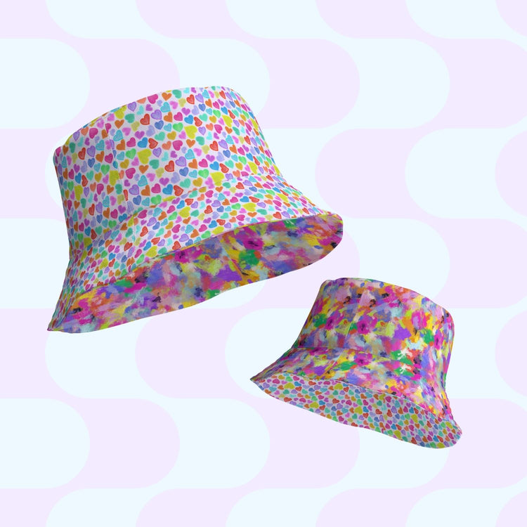 Two vibrant reversible bucket hats, the Heart Attack and Watercolour Rainbow by My Favourite Colour is Rainbow, are set against a pastel geometric backdrop. One exhibits a multicolored heart motif; the other flaunts an abstract splash pattern, making them perfect style companions available in various sizes.
