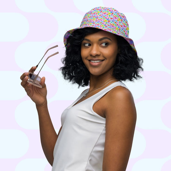 A curly-haired woman smiles while holding sunglasses, wearing the Heart Attack/Watercolour Rainbow Reversible Bucket Hat by My Favourite Colour is Rainbow, and a white sleeveless top, perfectly capturing the essence of Style BFF. A light geometric pattern completes the background.