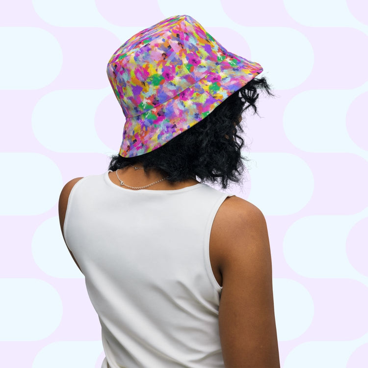 With curly black hair, someone wears a Heart Attack/Watercolour Rainbow Reversible Bucket Hat by My Favourite Colour is Rainbow and a sleeveless white top, embodying Style BFF vibes against a pastel geometric backdrop with their back facing the camera.