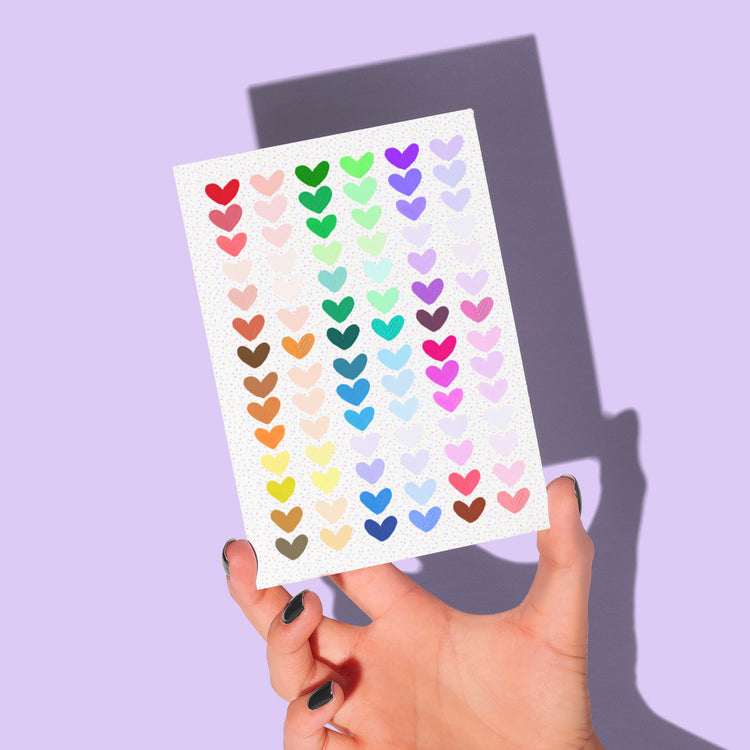 A hand with dark nail polish elegantly holds My Stores Heart Attack Greeting Card, adorned with vibrant heart-shaped stickers in a warm-to-cool gradient, casting shadows on a light purple background.