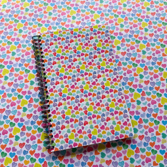 The spiral-bound Heart Attack Notebook by My Favourite Colour is Rainbow features a colourful heart pattern cover that blends seamlessly on a matching surface. Part of our vibrant collection, its hearts burst with creativity in shades of blue, pink, yellow, and green.