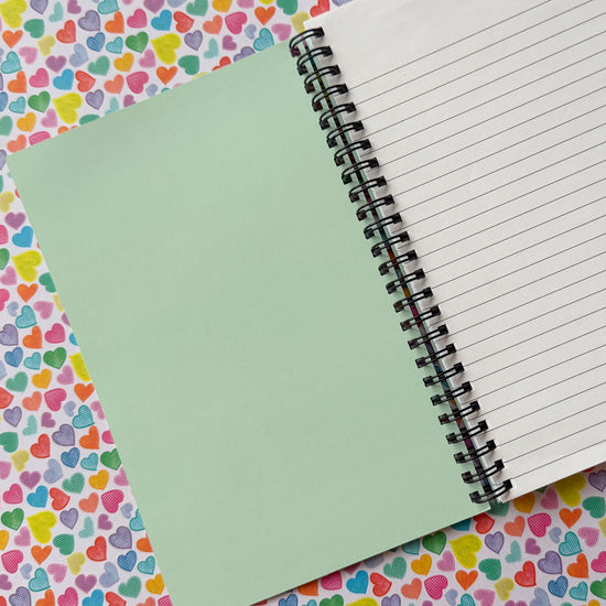 The inside cover of the Heart Attack notebook by My Favourite Colour is Rainbow, features a light green background. There is lined paper on the other side.  Set against a background of the same Heart Attack pattern as the notebook's covr.