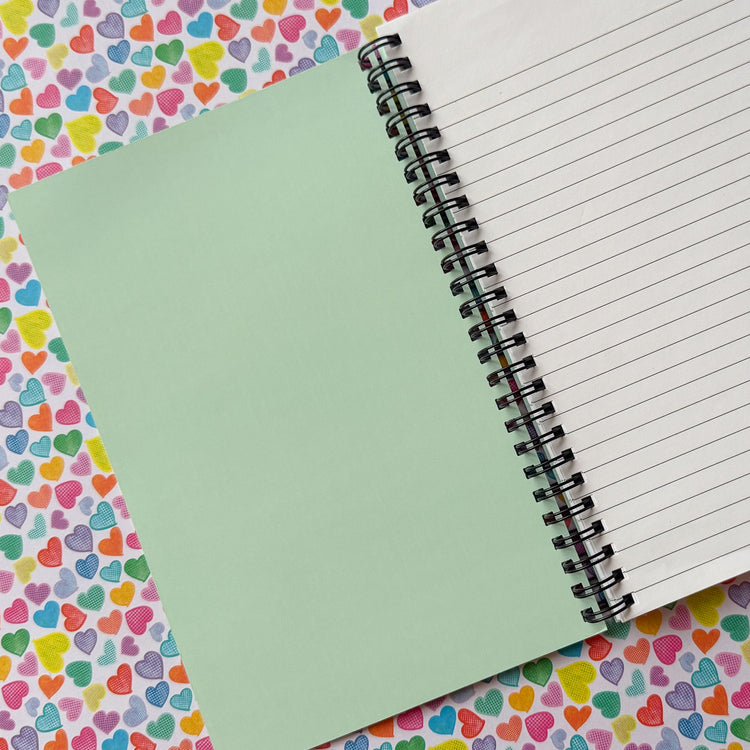 The inside cover of the Heart Attack notebook by My Favourite Colour is Rainbow, features a light green background. There is lined paper on the other side.  Set against a background of the same Heart Attack pattern as the notebook's covr.