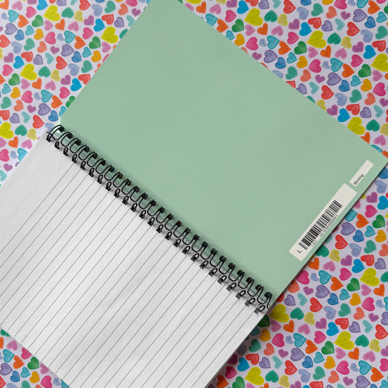 An open spiral Heart Attack Notebook by My Store, featuring lined pages and a green cover with a barcode, rests on a heart-patterned backdrop, sparking creativity as part of your vibrant stationery collection.