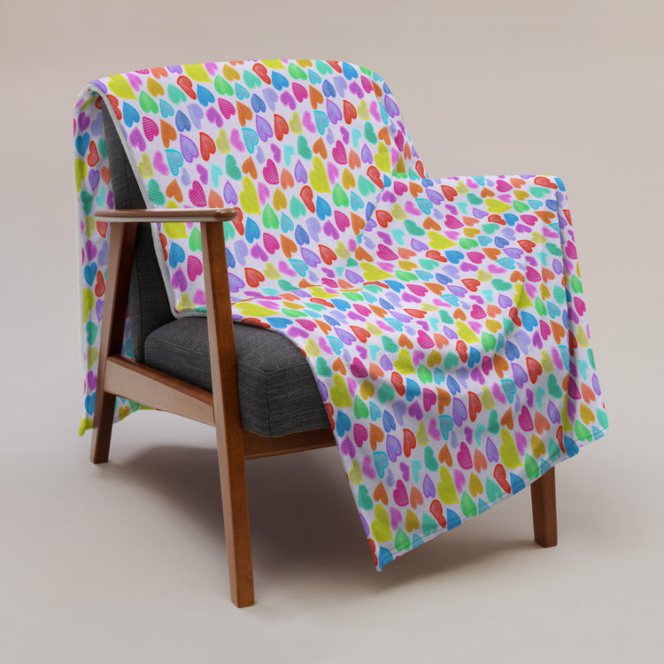 A wooden armchair with a gray cushion is adorned with the My Favourite Colour is Rainbows Heart Attack Throw Blanket, featuring vibrant multicolored overlapping heart shapes on soft fabric, in a minimal, neutral-toned setting.