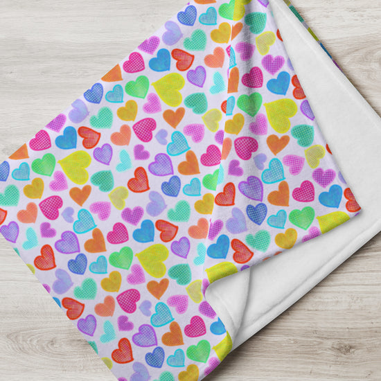 The Heart Attack Throw Blanket by My Favourite Colour is Rainbow features a vibrant multicolored heart design on a light background in blue, green, yellow, orange, pink, and purple shades. Its soft fabric elegantly drapes over the light wooden surface.
