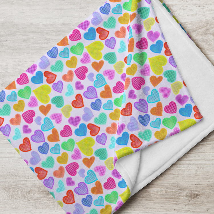 The Heart Attack Throw Blanket by My Favourite Colour is Rainbow features a vibrant multicolored heart design on a light background in blue, green, yellow, orange, pink, and purple shades. Its soft fabric elegantly drapes over the light wooden surface.