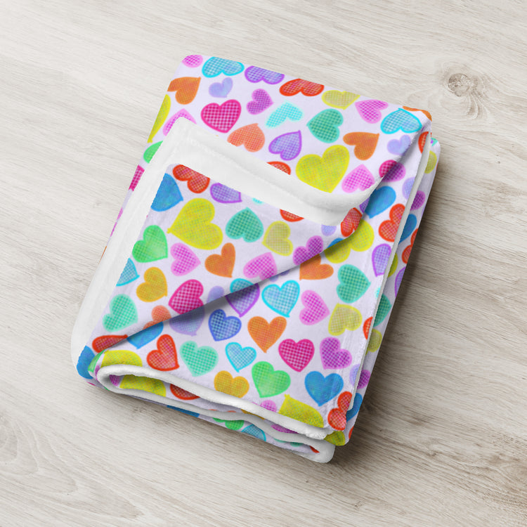 A Heart Attack Throw Blanket by My Favourite Colour is Rainbow, folded on a light wooden surface, displays a vibrant design with colorful hearts in pink, blue, green, yellow, orange, and purple.