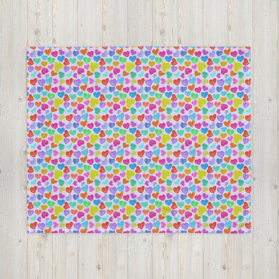 The Heart Attack Throw Blanket by My Favourite Colour is Rainbow features a lively multicolored heart design in pink, blue, green, orange, and purple on a light background, adding charm and character to the light wood floor.