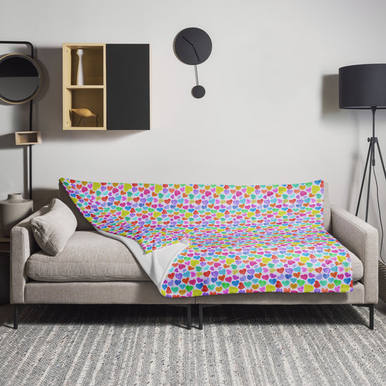A beige sofa is adorned with a My Favourite Colour is Rainbow Heart Attack Throw Blanket. A round black wall clock, black floor lamp, and decorative shelf items enhance the room, while a gray patterned rug adds texture to the cozy space.