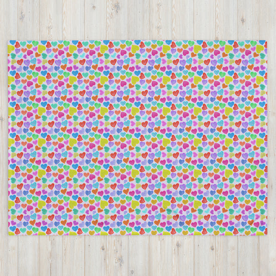 The Heart Attack Throw Blanket by My Favourite Colour is Rainbow features vibrant, overlapping hearts in pink, blue, green, and yellow against a light wood texture background—ideal for playful decor.