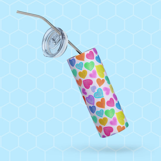 The Heart Attack Stainless Steel Tumbler from My Favourite Colour is Rainbow features a colorful heart pattern and includes a transparent lid with a stainless steel straw, displayed on a light blue honeycomb-patterned background.