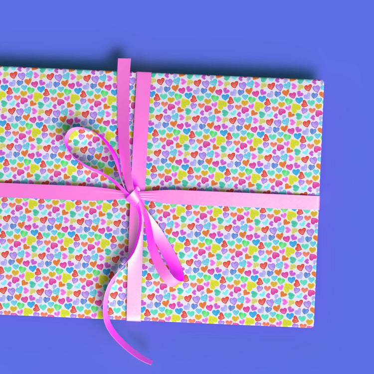 A rectangular gift box is wrapped in My Favourite Colour is Rainbows Heart Attack Wrapping Paper Sheets, featuring vibrant hearts and a pink ribbon. With a solid blue background, it’s perfect for eco-friendly gifting.