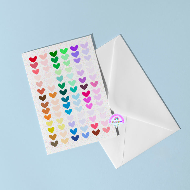 The Heart Attack Greeting Card by My Store features rows of colorful heart stickers on a bright card, set atop an open white envelope against a light blue background, with a charming round Rainbow sticker peeking from the edge.