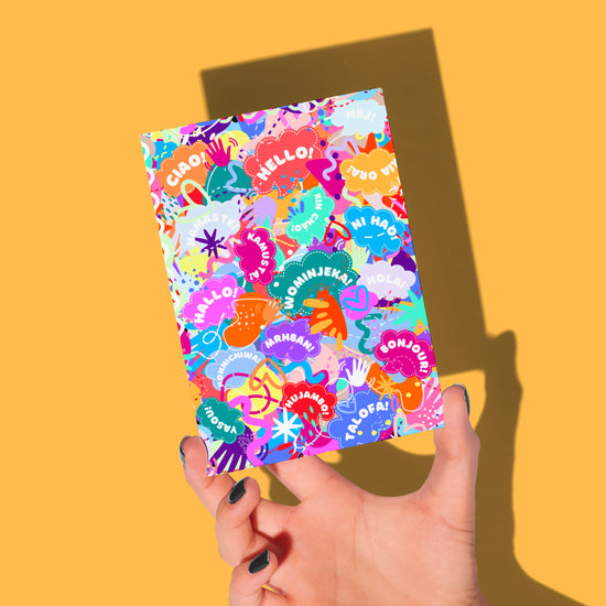 In a vibrant display, painted nails hold the Hello! Wominjeka! Greeting Card by My Store against an orange backdrop. This lively design showcases speech bubbles with greetings like Hello, Bonjour, and Hola, amidst cheerful doodles, sure to brighten anyones day.