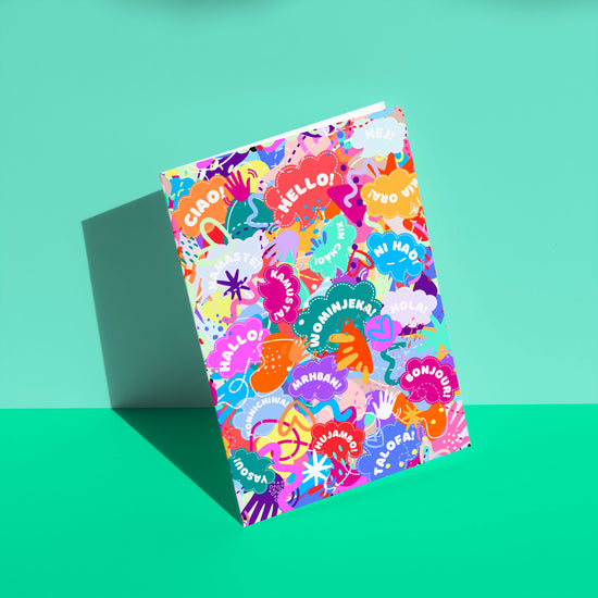 The Hello! Wominjeka! Greeting Card by My Store features a bold design with various greetings like Hello and Ciao, surrounded by abstract shapes. This vibrant card, angled against a teal background, radiates joyous diversity, sure to brighten anyones day.