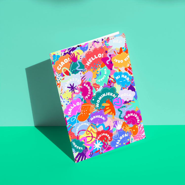 The Hello! Wominjeka! Greeting Card by My Store features a bold design with various greetings like Hello and Ciao, surrounded by abstract shapes. This vibrant card, angled against a teal background, radiates joyous diversity, sure to brighten anyones day.