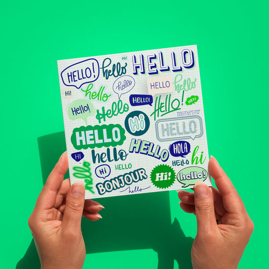 Two hands hold the Say Hello Greeting Card from My Favourite Colour is Rainbow, which features a green and blue collage of "hello: in different fonts and languages like hi, bonjour, and hola. It is shown against a green backgound.