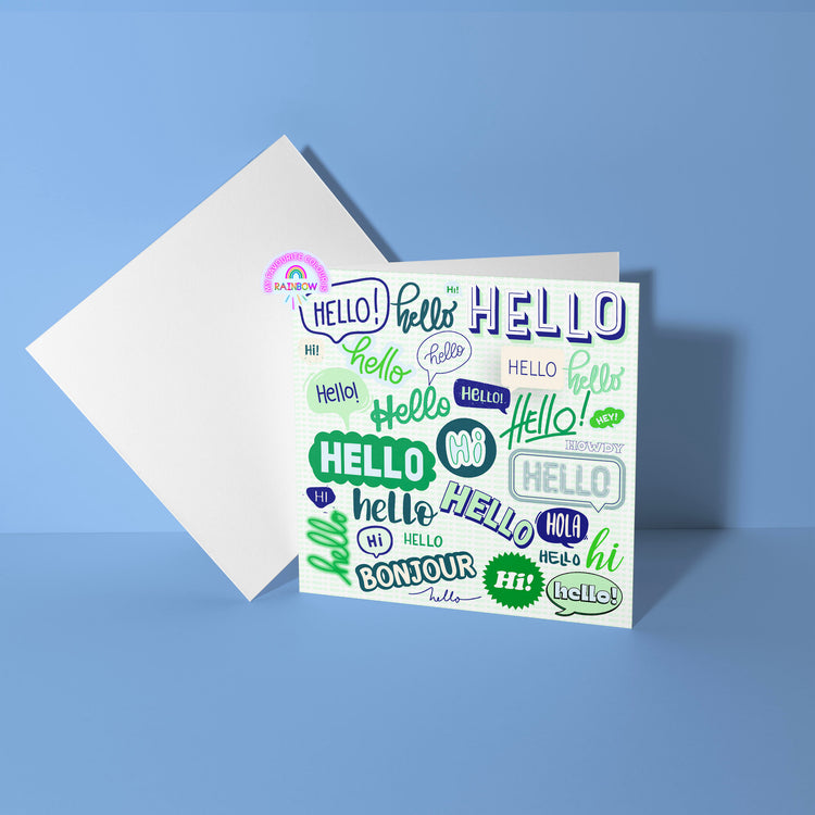 The Say Hello Greeting Card from My Favourite Colour is Rainbow features a colourful collage of hello in different fonts and languages like hi, bonjour, and hola. It is styled in blue and shades of green on a patterned white background.  It rests open on a blue surface with a white envelope behind it.