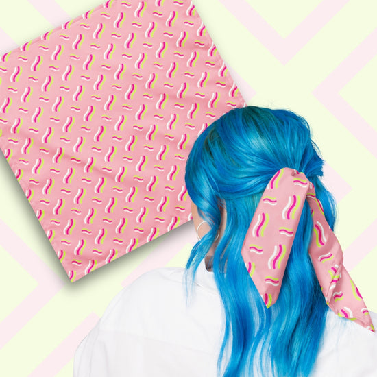 Two views of the 'Iced Vovo' Bandana by My Favourite Colour is Rainbow.  At top left, a flat lay of the bandana.  At bottom right, a person with vibrant blue hair is seen from behind, wearing the bandana in her hair.  The bandana features a pink background with white, hot pink and lime green wavy lines suggestive of the iconic Iced Vovo biscuit.