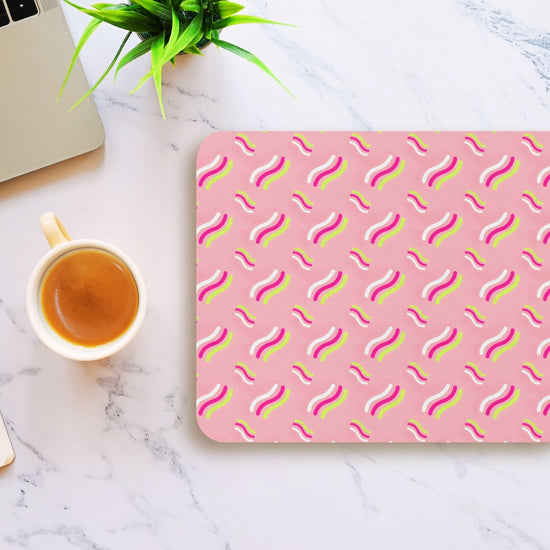 On a marble table sits a laptop, a cup of coffee, and an Iced Vovo Mouse Pad by My Favourite Colour is Rainbow, featuring a wavy pattern in white, yellow, and pink. A small green plant adds a touch of nature to the setup.