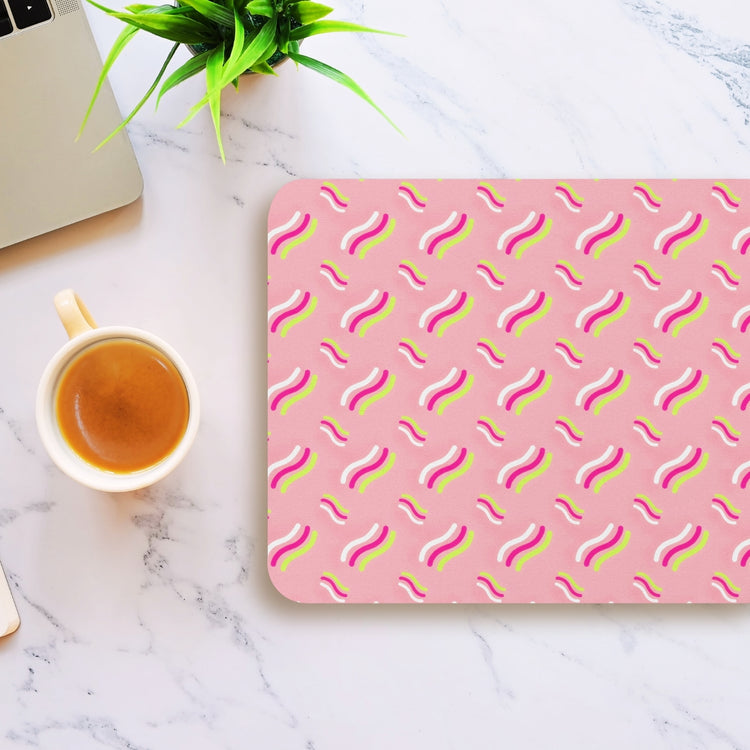 On a marble table sits a laptop, a cup of coffee, and an Iced Vovo Mouse Pad by My Favourite Colour is Rainbow, featuring a wavy pattern in white, yellow, and pink. A small green plant adds a touch of nature to the setup.