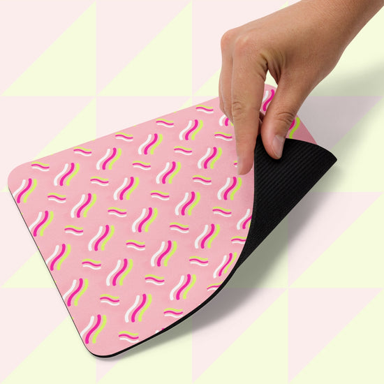 A hand lifts a corner of the Iced Vovo Mouse Pad by My Favourite Colour is Rainbow, showcasing its colorful abstract wavy pattern with a black underside. The background highlights a geometric design in pale green and white.