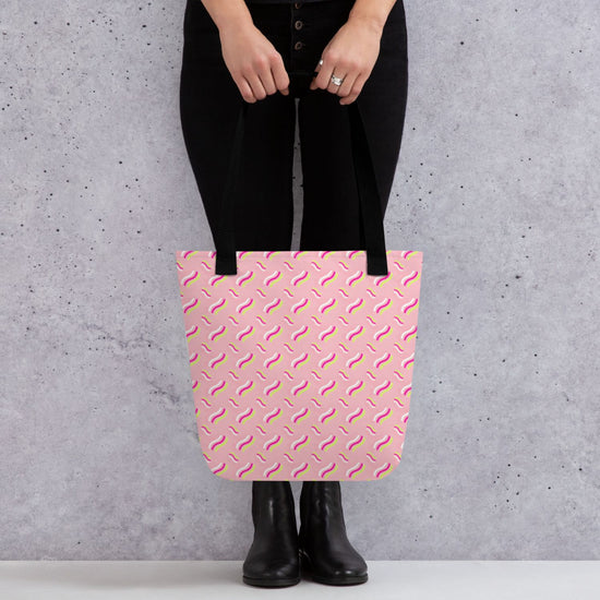 A person stands against a textured gray wall, wearing black pants and boots, holding the spacious Iced Vovo Tote Bag from My Favourite Colour is Rainbow. The pink bag features an abstract pattern reminiscent of the famous biscuit with white, pink and lime green wavy line patterns.