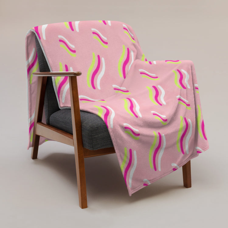 A dark gray cushioned mid-century modern armchair is draped with the Iced Vovo Throw Blanket by My Favourite Colour is Rainbow, featuring vibrant wavy stripes in white, pink, and lime green on a neutral background.