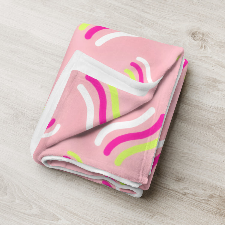 The Iced Vovo Throw Blanket from My Favourite Colour is Rainbow showcases a pink background with white, green, and pink curved lines. Its soft texture and vibrant design make it a perfect addition to any modern space, laying beautifully on a light wooden surface.