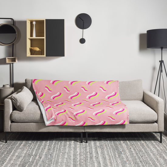 A modern living room features a beige sofa with the Iced Vovo Throw Blanket by My Favourite Colour is Rainbow, showcasing vibrant wavy patterns. A black wall clock, geometric shelves, and a tall lamp adorn the white wall, while a patterned rug adds warmth to the floor.