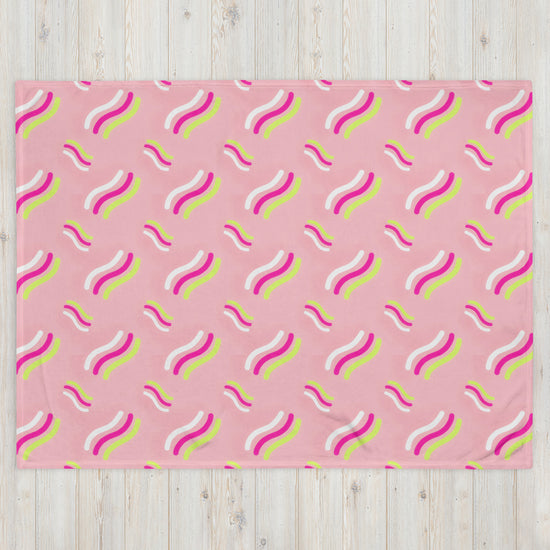 The Iced Vovo Throw Blanket by My Favourite Colour is Rainbow showcases a playful pattern of wavy lines in white, bright pink, and yellow on a light wooden background.