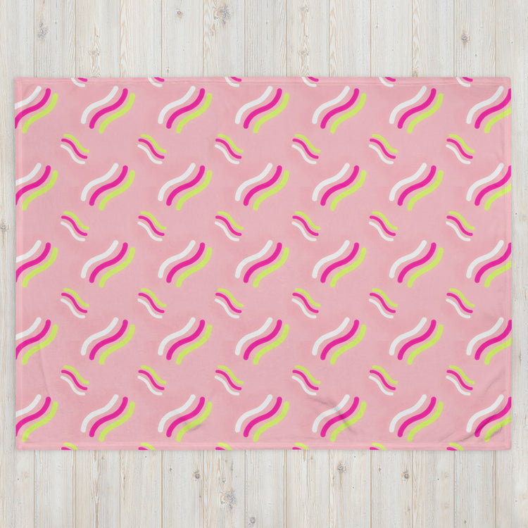 The Iced Vovo Throw Blanket by My Favourite Colour is Rainbow showcases a playful pattern of wavy lines in white, bright pink, and yellow on a light wooden background.