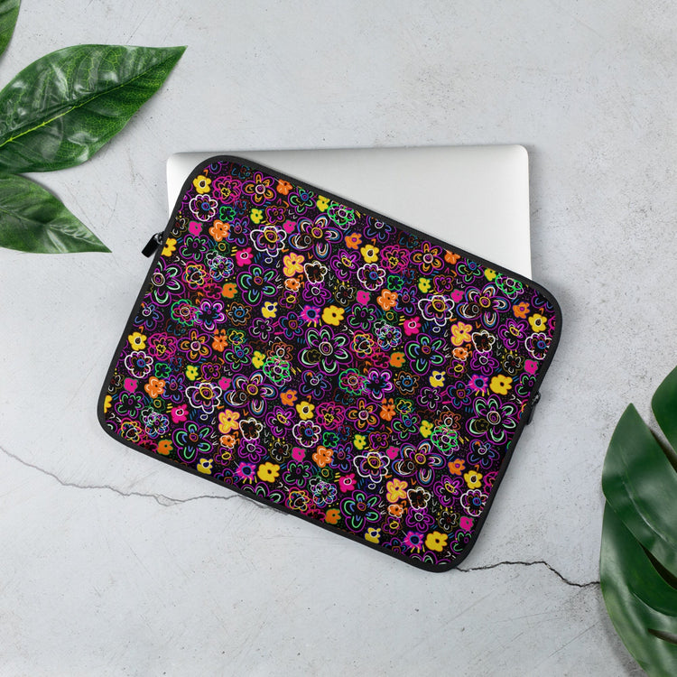 A laptop partially rests in the 13 inch 'In the Midnight Garden Laptop Sleeve' by My Favourite Colour is Rainbow.  The design features a vibrant floral design on a dark background.  It sits on a light grey surface surrounded by lush green leaves.